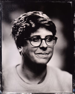 Author image taken in 2019, in tintype style of photograph. Eley is seated and bespectacled with short hair and an unsure expression.